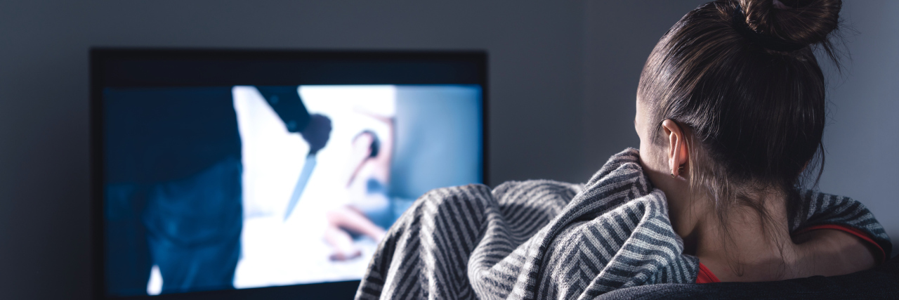How Watching Horror Movies Actually Helped Me Face My Anxiety A young white woman sitting on the couch hiding under a blanket, watching a horror movie