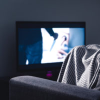 A young white woman sitting on the couch hiding under a blanket, watching a horror movie