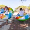 two children with a mask on their face and glasses walking through a pine forest campground with a colorful inflatable mattress wheel in their hands to enjoy a vacation in the midst of the covid 19 coronavirus pandemic