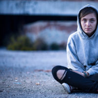 A young white female sitting criss-cross in the street with a hoodie on, serious facial expression