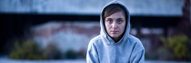 A young white female sitting criss-cross in the street with a hoodie on, serious facial expression