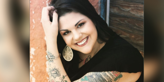 Photo of the author, a young white woman smiling, with a hand pushing her hair back, tattooed arm