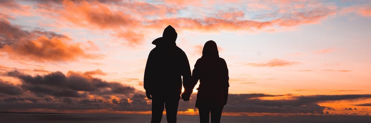 Silhouette of couple holding hands looking out over water and pink clouds at sunset
