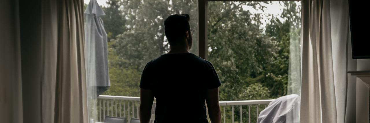 Why Saying 'It Could Be Worse' Isn't Helpful For Mental Illness photo of man looking out of a large window silhouetted against trees