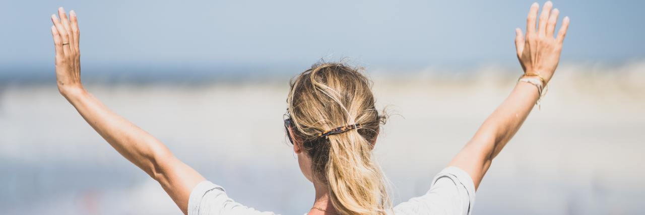 When Trauma and Chronic Illness Make You Hate Your Body Woman looking out over ocean and raising her hands in the air