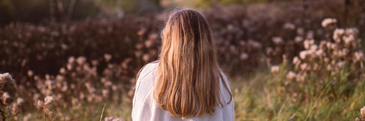 What I've Learned From Chronic Illness Back of a young woman in a white shirt with long blonde hair standing outside in a field of flowers