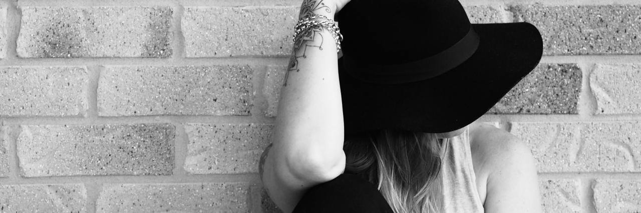 Why You Shouldn't Tell Someone With Depression to 'Think Positively' black and white photo of a woman against a brick wall with a black hat covering her face