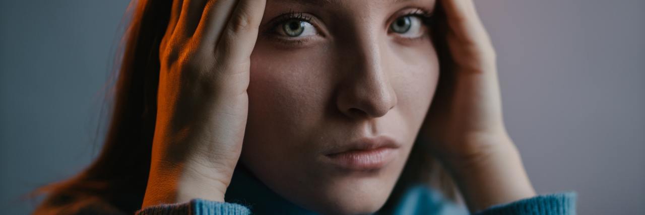 Signs You're Living With Toxic Shame photo of a woman looking into the camera with concern and sadness, hands at either side of her face