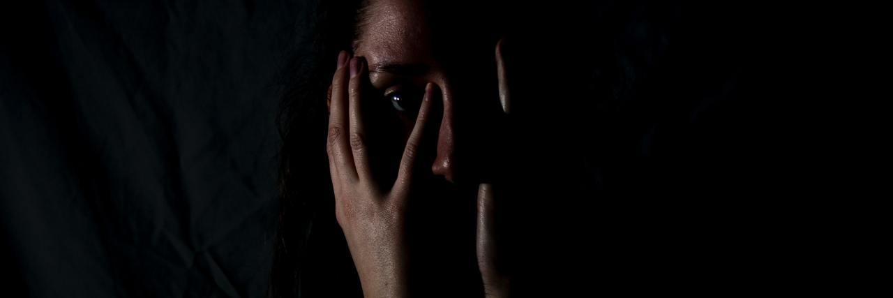 photo of a woman in darkness covering part of her face and mouth with her hands, looking into comera