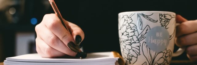 Photo of woman's hand writing in book next to a cup of coffee