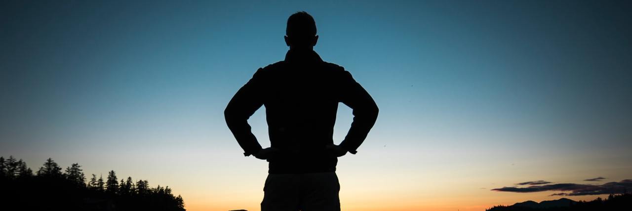 The back of a man standing with his hands on his hips, strong, overlooking a lake at sunset