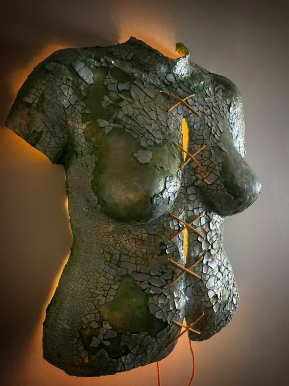 Body cast sculpture "Broken Body": torso in sea green color, covered in shining, cracked silvery pieces. String tied down the middle in a series making an "X" shape lit from behind