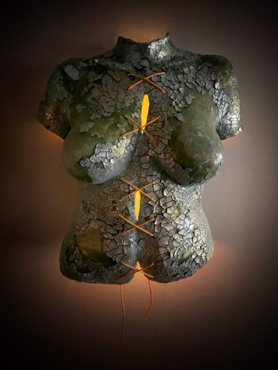 Body cast sculpture "Broken Body": torso in sea green color, covered in shining, cracked silvery pieces. String tied down the middle in a series making an "X" shape lit from behind