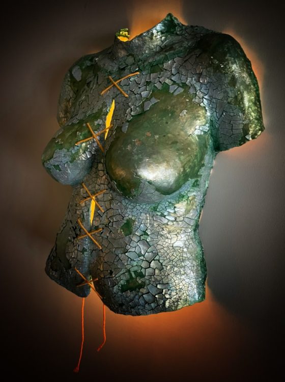 Body cast sculpture "Broken Body": torso in sea green color, covered in shining, cracked silvery pieces. String tied down the middle in a series making an "X" shape lit from behind