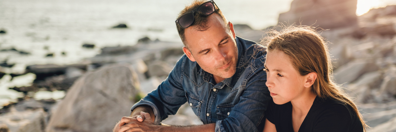 How I'm Learning to Support My Daughter With PTSD A father and teen daughter sitting on a rocky beach by the sea, father looking at daughter who has a serious facial expression