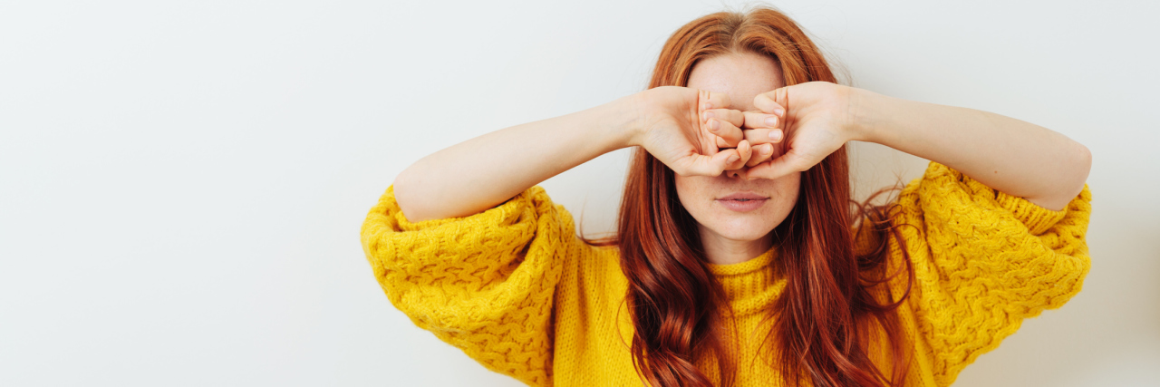 A young white woman with long red hair in a yellow sweater covering her eyes
