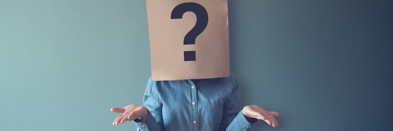 Woman in a denim button down shirt with a brown paper bag over head that has a question mark drawn on it