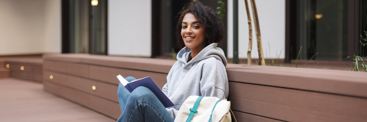 A young Black woman sitting against a wall in a grey sweatshirt with her backpack next to her, smiling