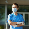 A white woman in scrubs standing outside a hospital in gloves and a mask, looking off into the distance