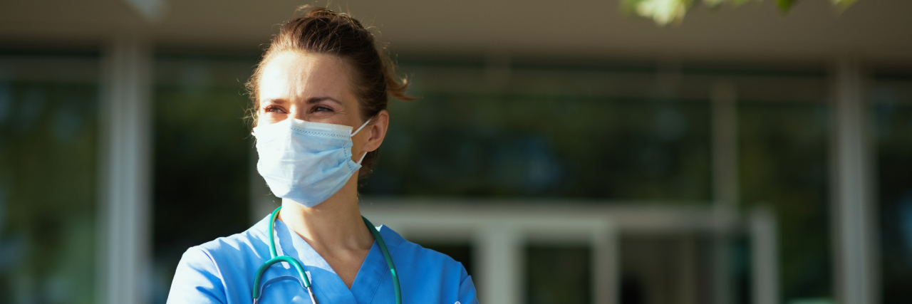 Battling Mental Health Stigma as a Nurse With Bipolar Disorder A white woman in scrubs standing outside a hospital in gloves and a mask, looking off into the distance
