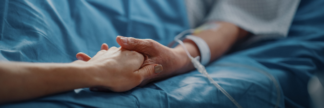 When We Say We'll 'Be There' When a Loved One Is Dying An older white woman lying in a hospital bed with an IV hooked up to her arm, and another woman's hand holding hers