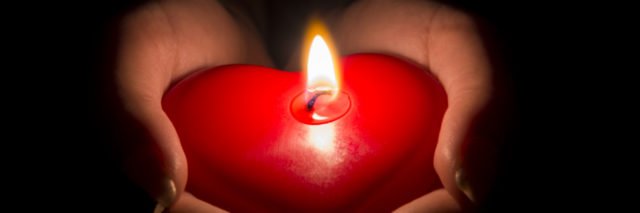 Woman's hands holding a heart shaped candle in the dark.