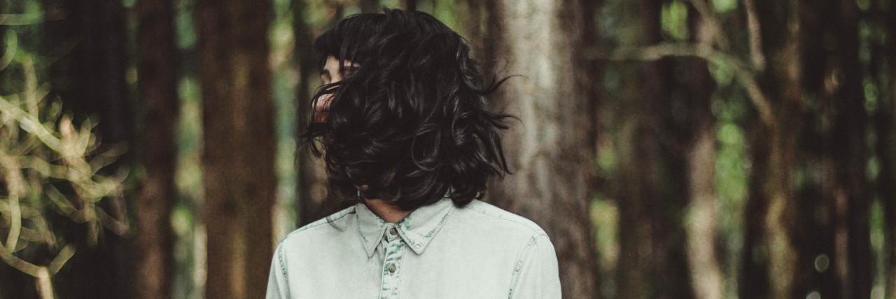 Chronic Illness Treatment as a Survivor of Medical Child Abuse A young white woman with dark hair covering her face, standing in a forest