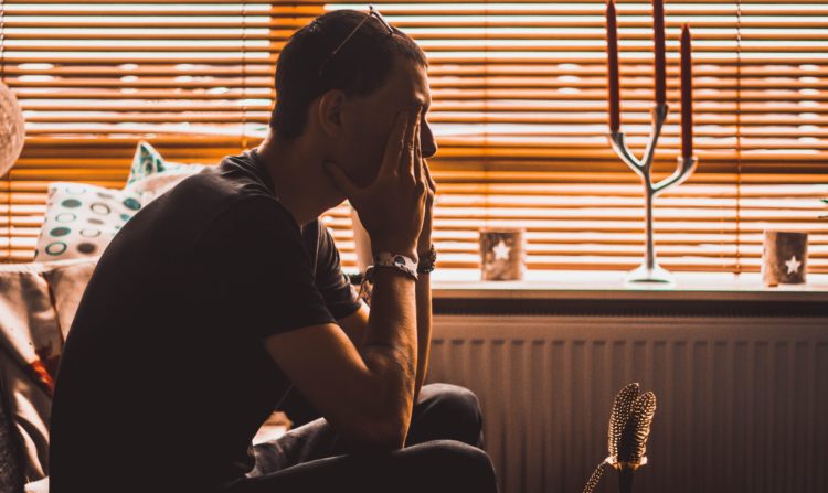 photo of a man sitting on a bed in front of window with his hands covering his face