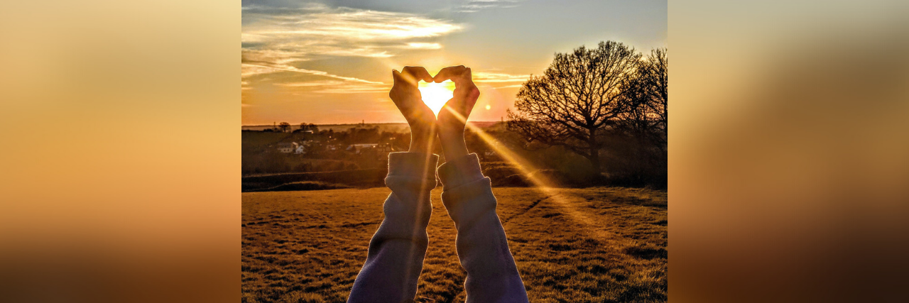 Person holding up hands in a heart shape with sun coming through them at sunset