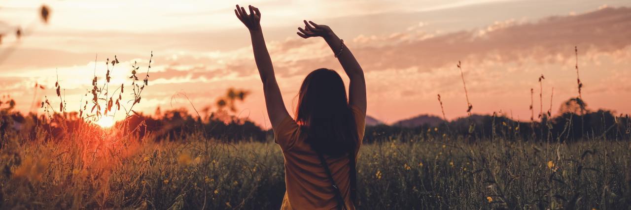 A woman at sunset in a peaceful field with her hand raised above her head.