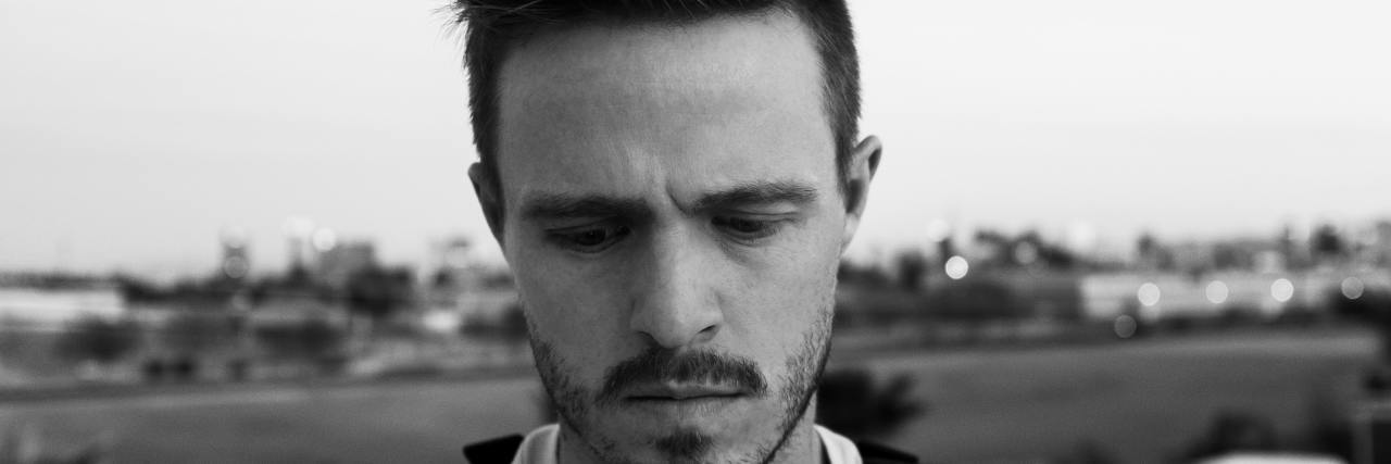 Black and white photo of a young, white man with a backpack on, looking down with a serious expression, short spiky hair