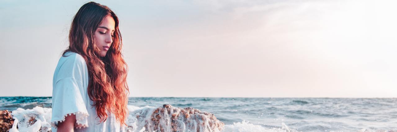 A young woman with long hair standing in an ocean by rocks with a white t-shirt on looking down