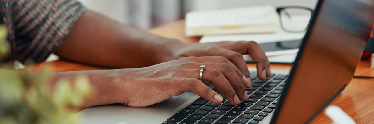 Black woman's hands typing on laptop on desk by pad of paper, glasses and pens