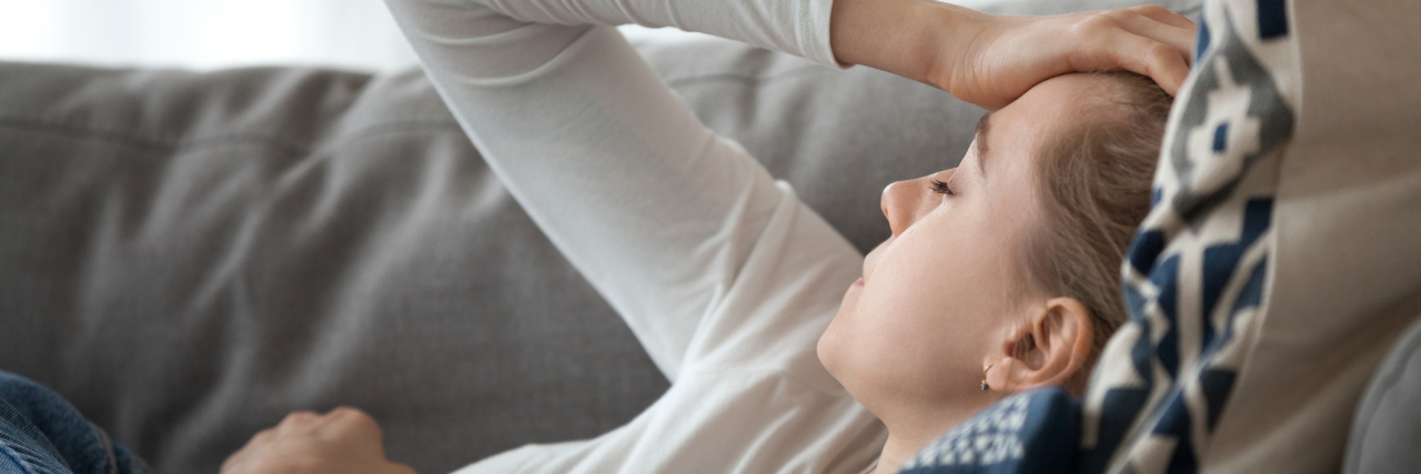 COVID-19 Long-Haulers Should Qualify for Disability Benefits A blonde woman with pale skin lying down on the couch with her hand on her head