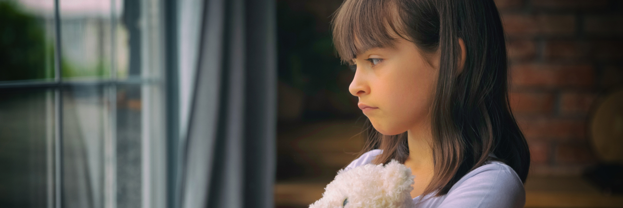 Sad little girl hugging toy, looking out window.