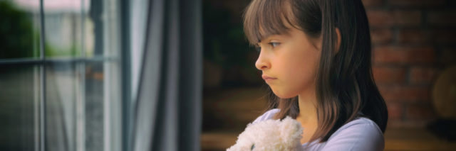 Sad little girl hugging toy, looking out window.