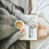 Photo of woman wearing a sweater, holding a book and cup of coffee while relaxing at home