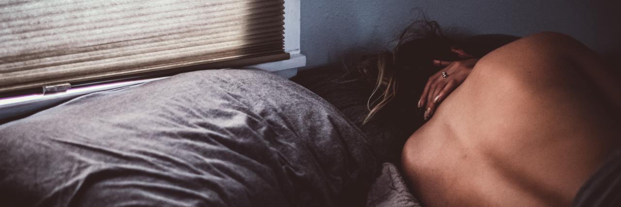 The Reality of a 'Typical' Day, Week and Year With Health Anxiety a young, white woman lying in bed facing the wall with a hand on her neck in pain