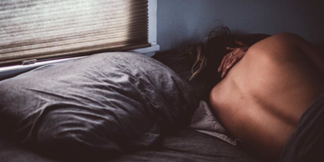 a young, white woman lying in bed facing the wall with a hand on her neck in pain