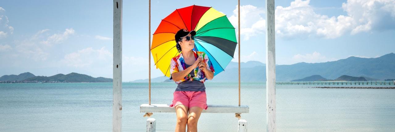 What Does It Mean to Be Neurodivergent? Photo of woman sitting on a swing holding a rainbow umbrella in front of the sea