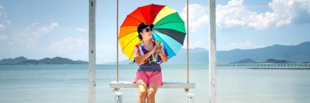 Photo of woman sitting on a swing holding a rainbow umbrella in front of the sea