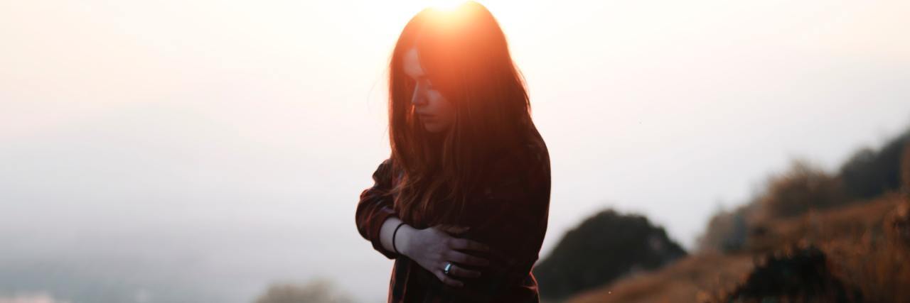 Just as I Was Discharged From the Psych Ward, I Found Out I Had Cancer A young woman standing on a hill in a red flannel shirt, touching her arm and looking down solemnly
