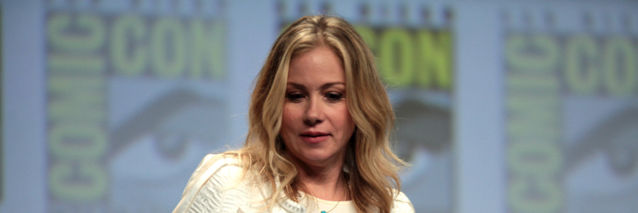 Christina Applegate standing at San Diego Comic Con at a panel. She's wearing a white blouse and some pretty gold necklaces.