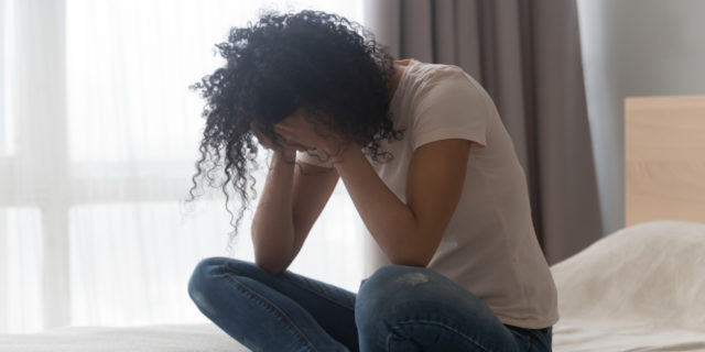 Young Black woman sitting on her bed crying, hands to her face