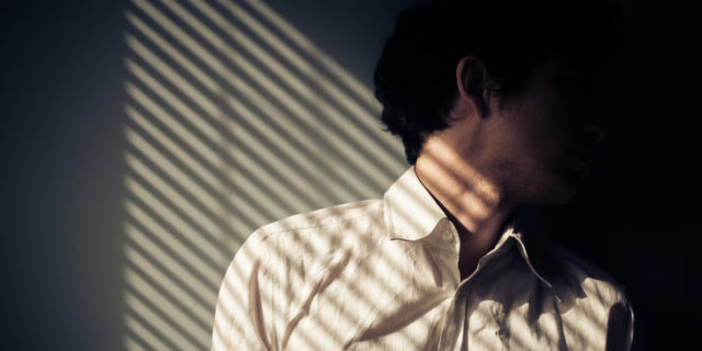 When the Summer Sun Is Not so Fun for Those With Chronic Illness Young man next to a window with shadows being cast from the blinds