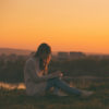 A young woman sitting by a lake at sunset writing in a journal