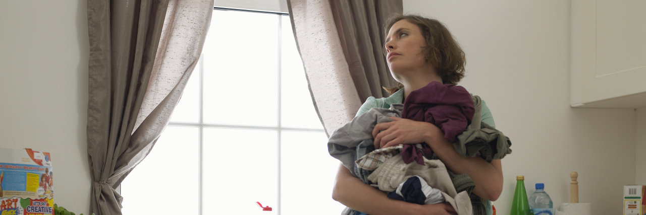 Dealing With the Daily Drudgery of Life With a Chronic Illness Woman holding laundry in a messy kitchen.