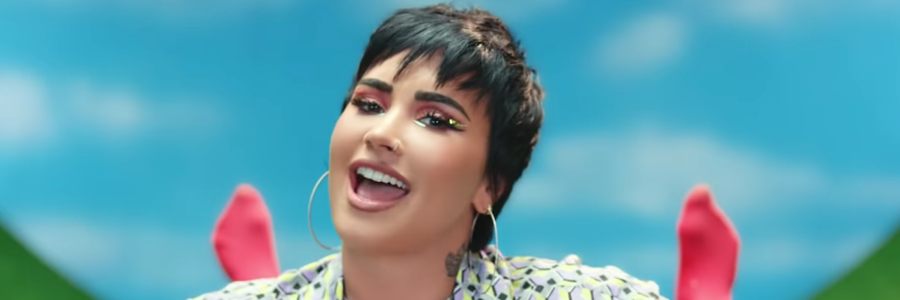 Why Demi Lovato's Song 'Melon Cake' Is a Great Eating Disorder Anthem Demi Lovato smiling in their new music video "Melon Cake." They have on a patterned shirt with a diamond choker, and a short hair cut. They're surrounded by a blue sky backdrop and upside down feet. They're in hand stands.