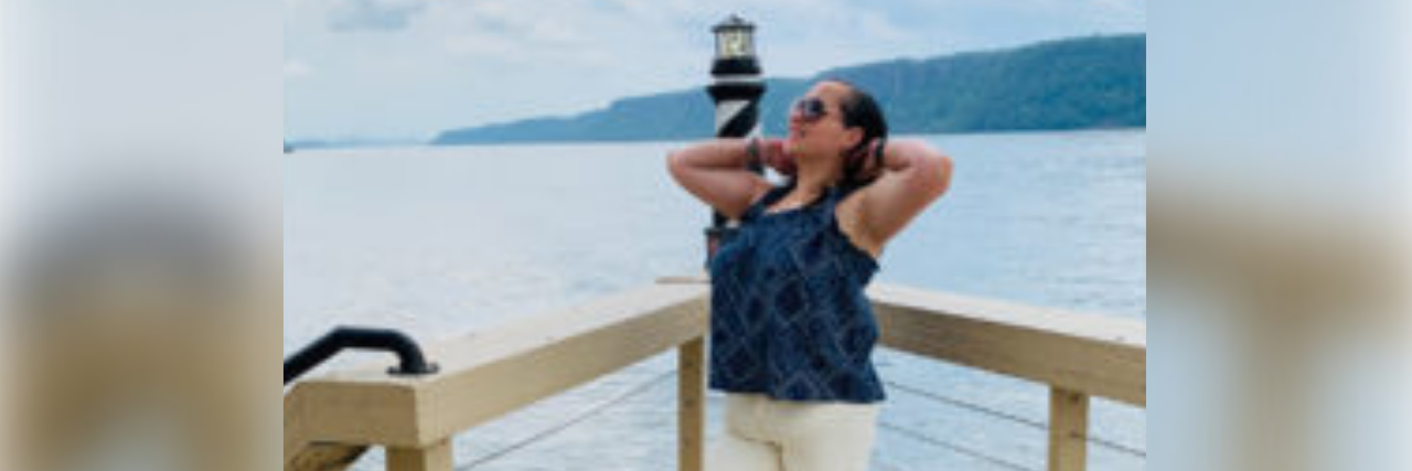 Finding a Work-Life Balance With Chronic Trauma and Fibromyalgia Author, a middle-aged white woman standing on a dock posing in front of a lighthouse