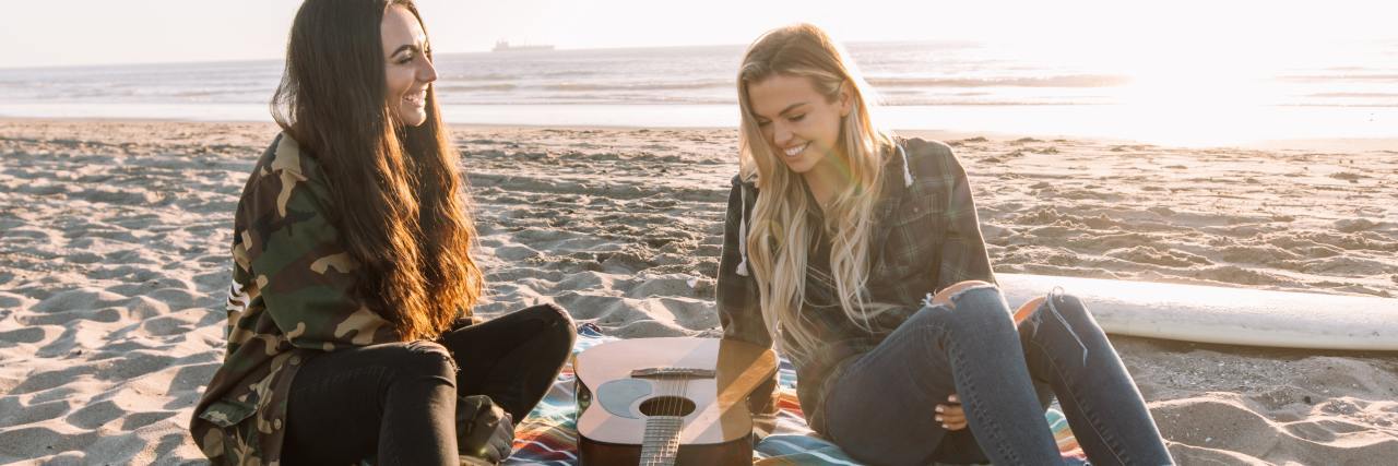 photo of two women sitting on a beach with a picnic and guitar